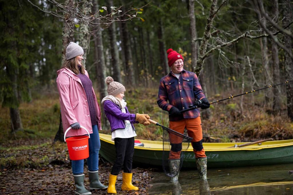 Kalassa metsälammen rannassa seisoskeleva hymyilevä ja naursekeleva kolmihenkinen perhe mukanaan virveli, haavi sekä Tokmannin punainen brändiämpäri.