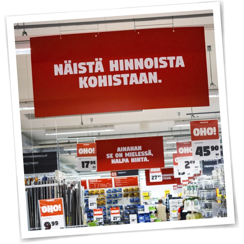 Tokmannin myymälän sisätila, jossa roikkuu punaisia hintakylttejä. Etualalla suuri kyltti, jossa lukee "Näistä hinnoista kohistaan." Taustalla näkyy hyllyrivejä ja muita tarjouksia korostavia kylttejä, kuten "Oho!" ja "Ainahan se on mielessä, halpa hinta."