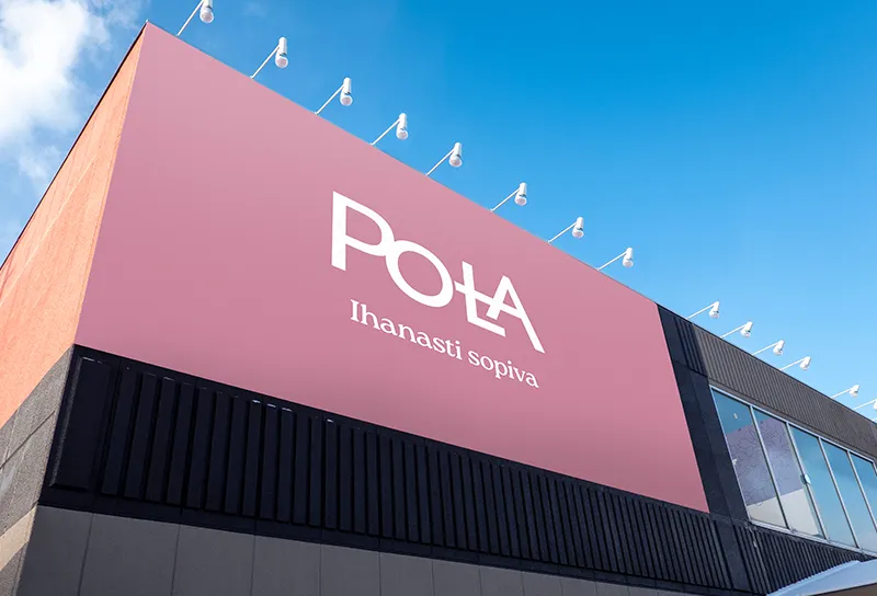 Vaaleanpunainen julkisivutaulu, jossa valkoinen, modernisti typografinen "POLA"-logo. Kirjaimessa L on poikkiviiva, joka yhdistää sen A-kirjaimeen luoden erottuvan visuaalisen ilmeen. Logon alla teksti "Ihanasti sopiva" täydentää brändiviestiä lempeällä ja kutsuvalla sävyllä. Tausta on selkeä ja taivas kirkas, mikä korostaa brändin raikkautta ja selkeyttä.
