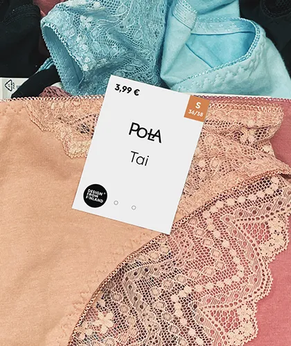 Naisten pitsialushousut vaalean persikan sävyssä esillä myymälässä. Tuotteen päällä on valkoinen hintalappu, jossa lukee “3,99 €”, “Pola”, “Tai” sekä koko “S 36/38”. Lapussa on myös “Design from Finland” -merkki. Taustalla näkyy muita alushousuja sinisen ja mustan sävyissä.