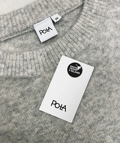 Vaaleanharmaa neulepusero, jossa resorillinen pyöreä pääntie. Neuleen sisäpuolella näkyy kokolappu (koko M) ja Pola-logolla varustettu merkki. Neuleen päällä lepää valkoinen hintalappu, jossa Pola-logo ja musta Design from Finland -merkintä.