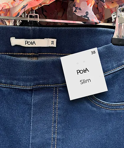 Naisten siniset slim-malliset farkut esillä henkarissa. Farkkujen vyötäröllä näkyy Pola-logolla varustettu kokolappu (koko 38) sekä erillinen hintalappu, jossa lukee “Pola”, “Slim” ja “38”. Taustalla näkyy vaaleanpunaisia ja oranssin sävyisiä kukkakuosisia vaatteita.