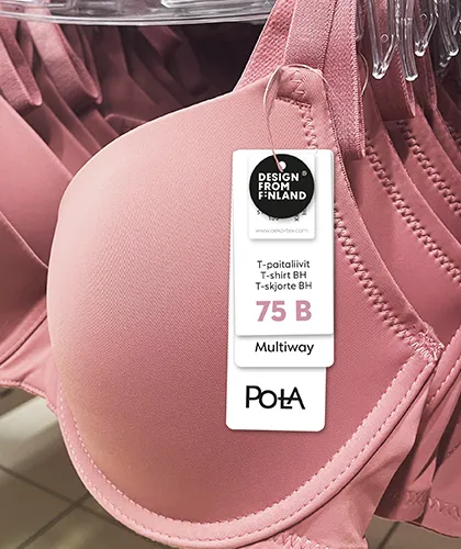 Vaaleanpunainen T-paitaliivi (rintaliivit) roikkumassa myymälän vaaterekissä. Tuotteessa on kiinnitettynä valkoinen hintalappu, jossa näkyy merkinnät: "DESIGN FROM FINLAND", koko "75B", tuotetyyppi "T-paitaliivit / T-shirt BH / T-skjorte BH", ja ominaisuus "Multiway". Alareunassa on Pola-tuotemerkin logo mustalla tekstillä. Taustalla näkyy useita päällekkäisiä samanlaisia rintaliivejä ripustettuna.
