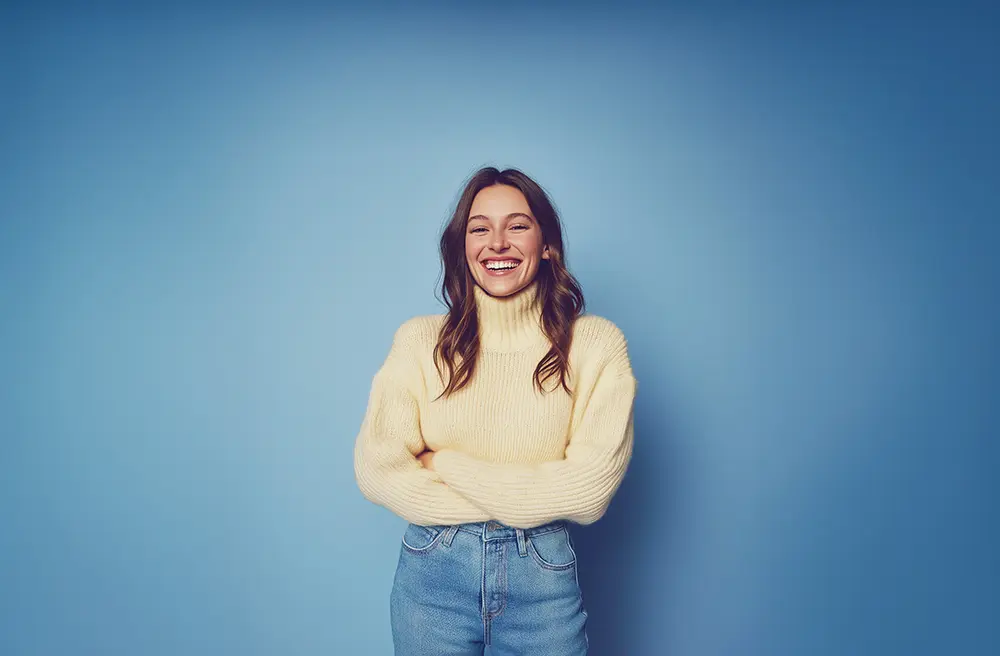 Naurava nainen seisoo sinistä taustaa vasten kädet ristissä. Hänellä on yllään vaaleankeltainen ribbineule ja siniset farkut. Kuvassa välittyy iloisuus, rentous ja itsevarmuus.