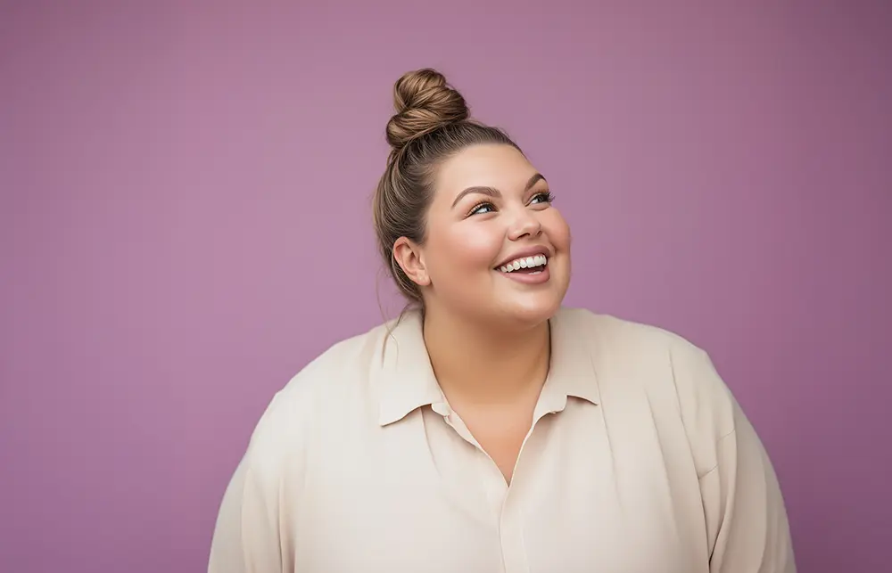 Iloinen nainen katsoo ylöspäin ja hymyilee leveästi vaaleanviolettia taustaa vasten. Hänellä on vaaleasävyinen pusero ja hiukset nutturalla. Kuvassa korostuvat positiivisuus ja itsevarmuus.