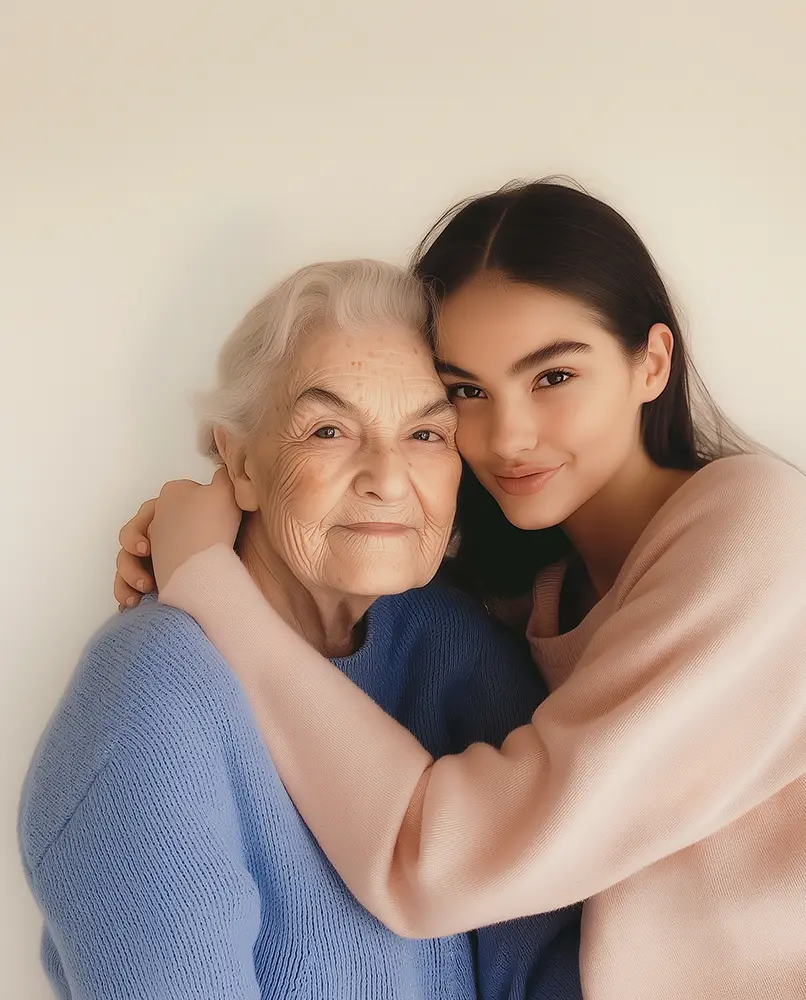 Alt-teksti: Nuori nainen halaa vanhempaa naista hellästi. Molemmat hymyilevät, nojaavat toisiinsa posket vastakkain ja katsovat suoraan kameraan. Vanhuksella on yllään sininen neule, nuoremmalla vaaleanpunainen. Tausta on vaalea ja neutraali. Kuvassa välittyy sukupolvien välinen lämpö ja läheisyys.