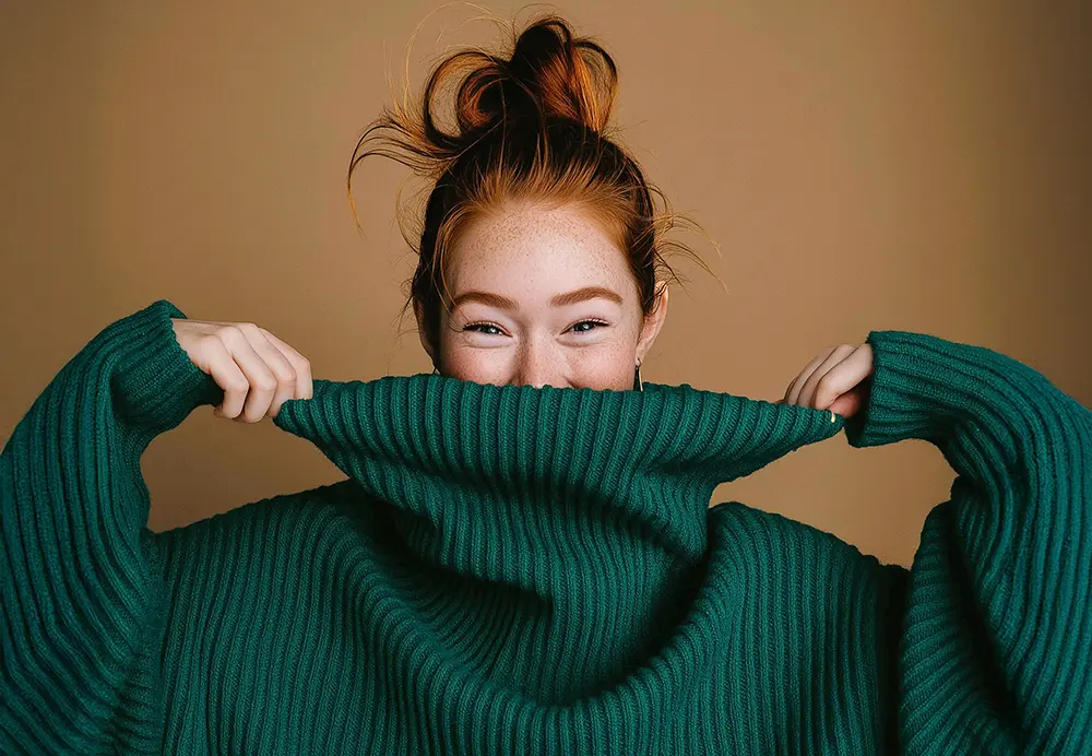Hymyilevä nainen vihreässä runsaan kokoisessa ribbineuleessa nostaa neuleen kaulusta kasvojensa eteen peittäen suunsa. Kuvan taustalla on lämpimän ruskean sävyinen seinä.