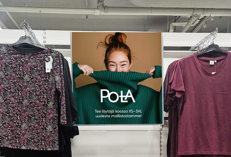 Mainosjuliste vaateosaston seinällä: nuori nainen katsoo kameraan vihreän poolopaidan kauluksen takaa iloisesti hymyillen. Julisteessa valkoinen Pola-logo ja teksti "Tee löytöjä koossa XS–3XL uudesta mallistostamme!" Julisteen molemmin puolin roikkuu vaatteita: vasemmalla kukkakuosinen ja musta vaate, oikealla viininpunainen t-paita. Taustalla näkyy myymälän kattoa ja putkistoa. Kuvassa korostuvat positiivinen tunnelma, helposti lähestyttävä brändi-ilme ja selkeä viestinnällinen kokonaisuus.