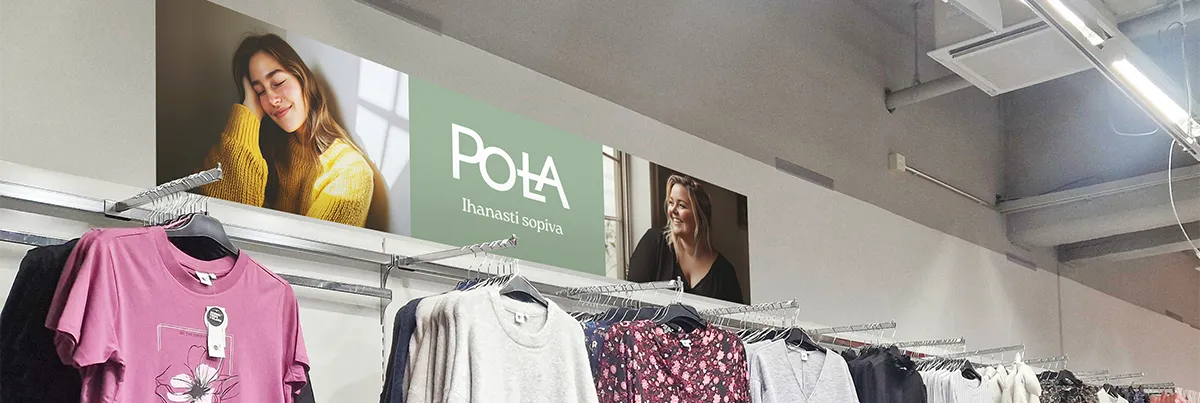 Vaateosaston yläpuolella seinään kiinnitetty pitkä, vaakasuuntainen mainos Pola-vaatemallistosta. Mainoksessa on keskellä vihreä taustapalkki, jossa valkoinen Pola-logo ja iskulause "Ihanasti sopiva". Molemmilla puolilla näkyy valokuvia iloisista naisista – vasemmalla nainen keltaisessa villapaidassa, oikealla nainen mustassa trikoopaidassa. Alapuolella roikkuu naisten vaatteita rekeissä, kuten neuleita ja kukkakuosisia puseroita. Mainos luo lämpimän ja kutsuvan tunnelman.