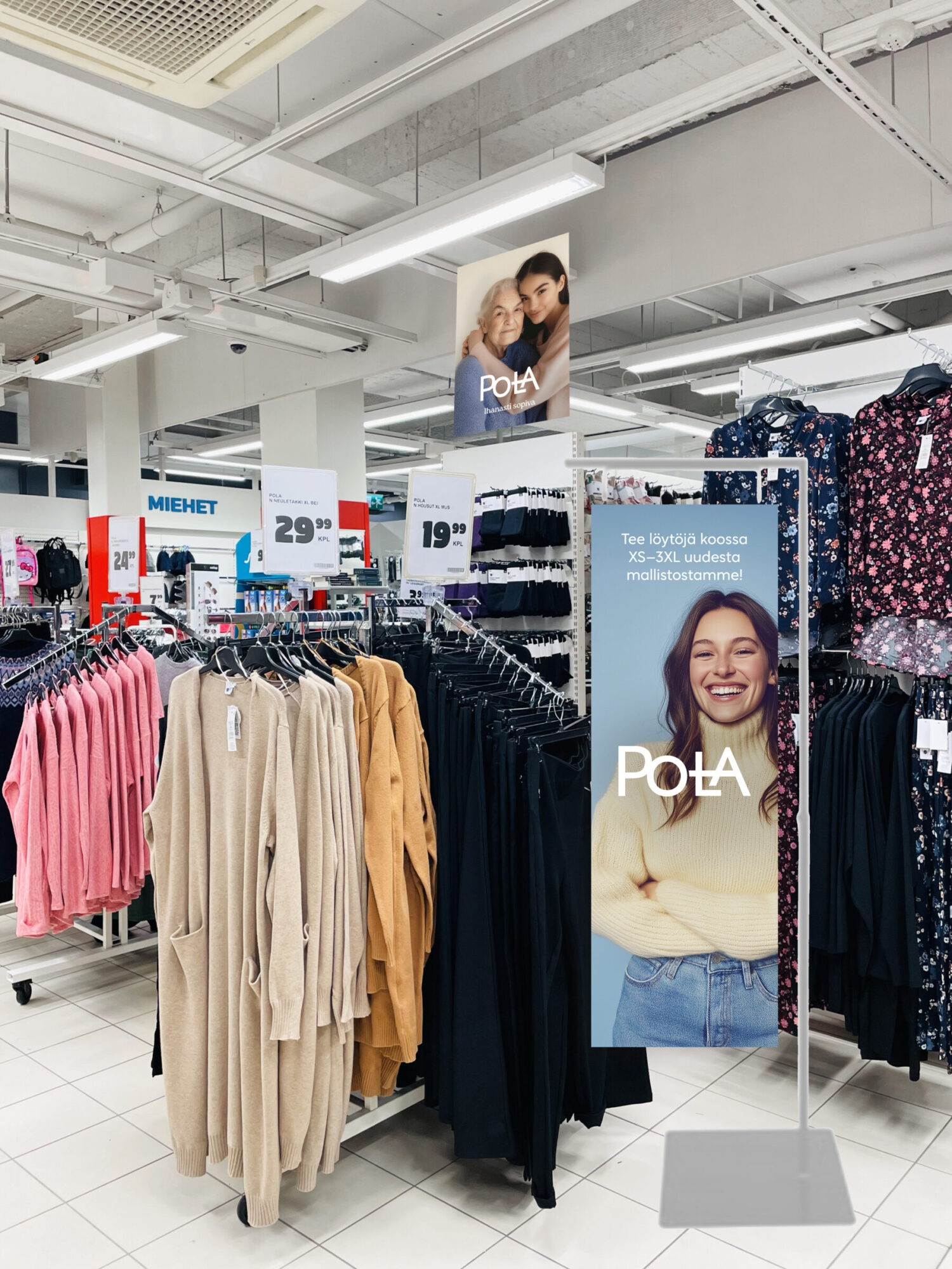 Myymäläympäristö, jossa Pola-vaatteet ovat esillä selkeissä vaaterekeissä. Etualalla vaaleita, kerman ja ruskean sävyisiä neuletakkeja, taustalla mustia ja kukallisia vaatteita. Keskellä lattiateline, jossa iloinen nainen ja teksti: "Tee löytöjä koossa XS–3XL uudesta mallistostamme!" sekä Pola-logo. Ylhäällä roikkuu lisäjulisteita, joissa samaa brändi-ilmettä. Hintakyltit mustavalkoisia, kirkkaalla fontilla. Visuaalisesti raikas, moderni ja helposti navigoitava ostoympäristö.