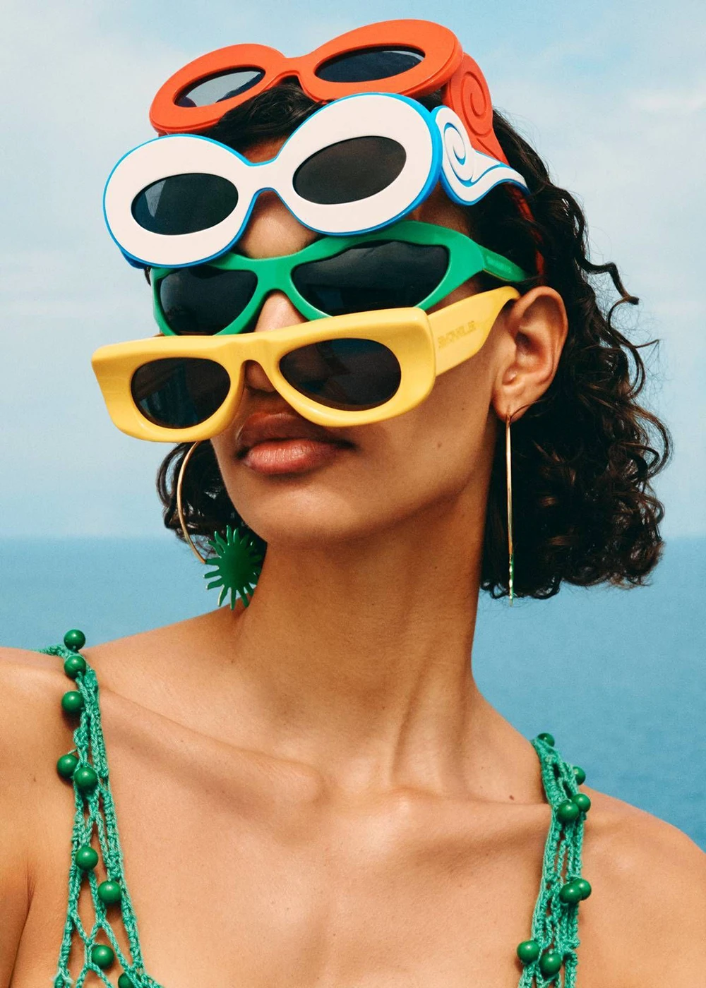 Product image of a woman dressed in a green bikini, wearing multiple pairs of colorful Miny sunglasses on her face in front of the sea