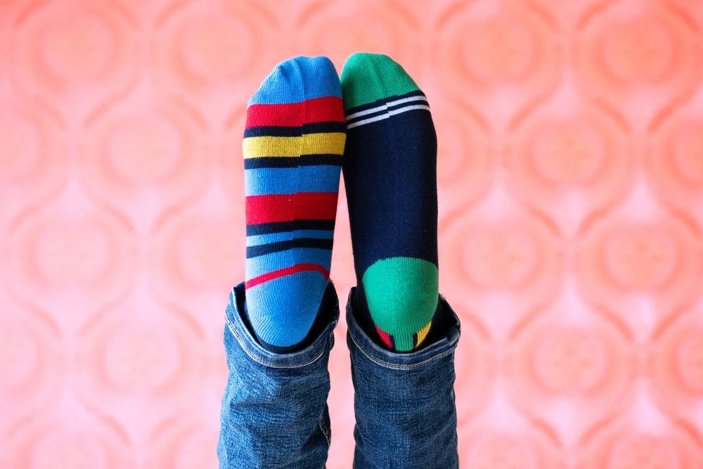 Product image of feet wearing mismatched colorful Miny socks with jeans against a pink patterned background