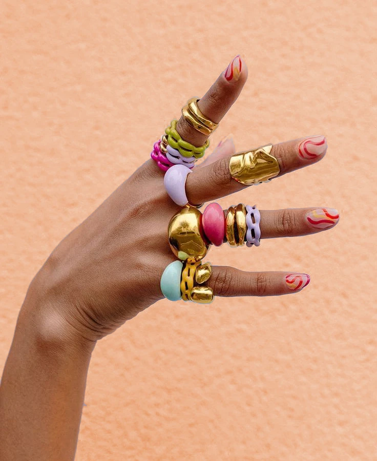 Product image of a hand wearing plenty of colorful Miny rings and decorated nails against a peach-toned background