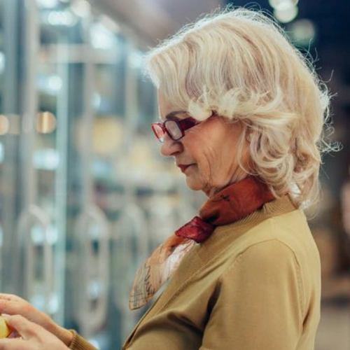 Brand persona Pirkko: a fair-haired woman of retirement age, wearing glasses, examining products on a store shelf.