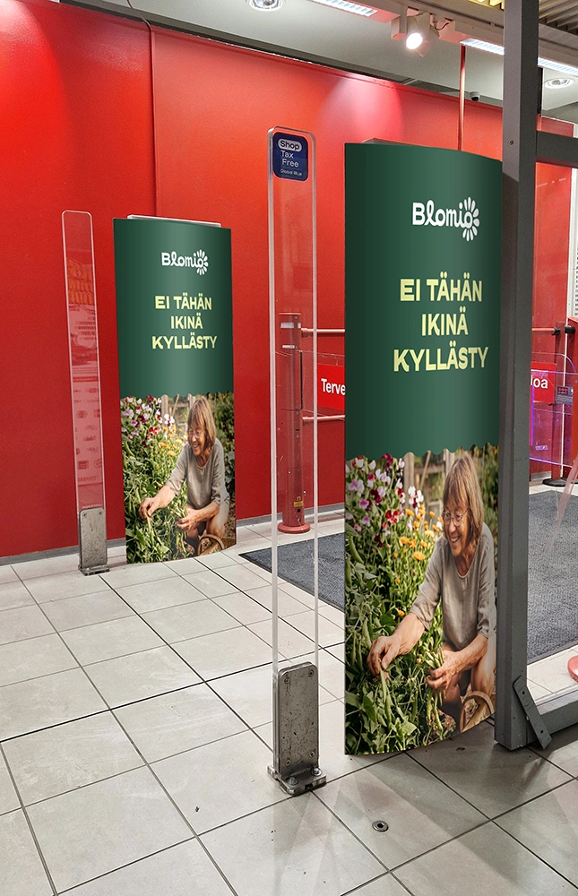 Myymälän sisäänkäynnin hälytysportteihin kiinnitetyt Blomio-mainokset ”Ei tähän ikinä kyllästy”, joissa kuva naisesta kukkapenkin ääressä.