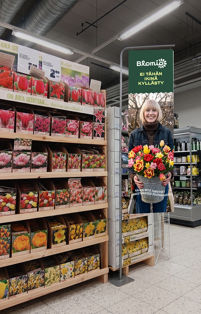 Myymälän siemenpussihylly ja sen vieressä Blomio-mainosteline ”Ei tähän ikinä kyllästy”, jossa nainen pitelee tulppaanikimppua.