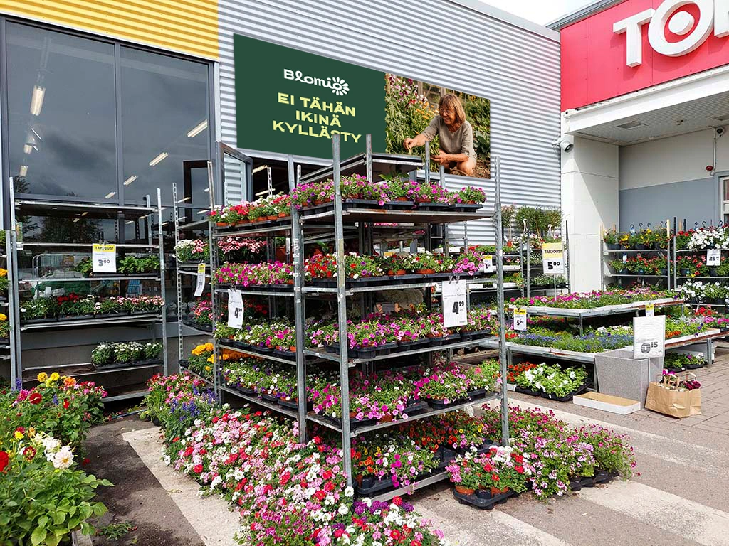 Myymälän pihan kesäkukkamyynti, ja taustalla seinässä Blomio-mainosteksti ”Ei tähän ikinä kyllästy” ja kuva naisesta puutarhassa.