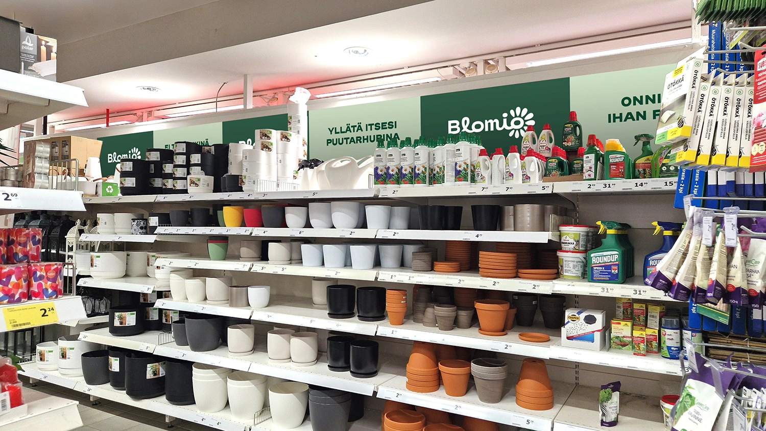 Myymälän puutarhahylly, jonka ylimmän hyllyn takaseinällä on vihreä Blomio-mainosnauha, jossa vuorottelee Blomion logo, sekä mainostekstejä kuten "Yllätä itsesi puutarhurina".
